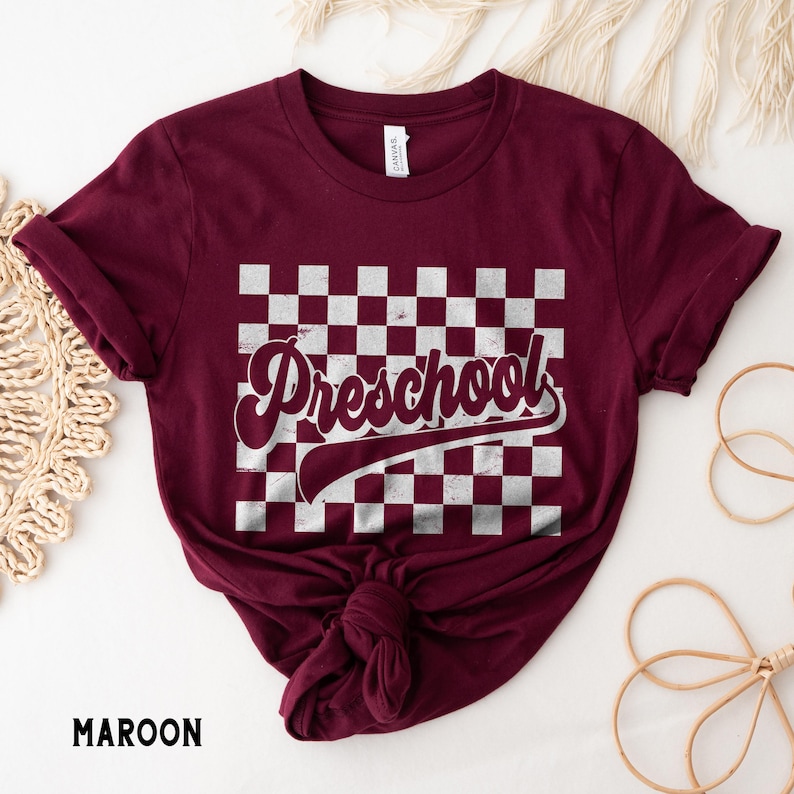 Pu&ograve; includere: T-shirt bordeaux con motivo a scacchi bianchi e grigi e la scritta "Preschool" in un font retr&ograve;. La maglietta &egrave; annodata in basso. La parola "MAROON" &egrave; stampata in basso a sinistra.