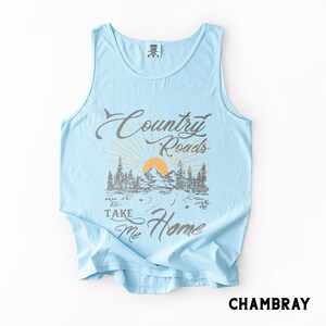 K&ouml;nnte beinhalten: Hellblaues Tanktop mit dem Text "Country Roads Take Me Home" in einer grau-orangenen Grafik. Die Grafik zeigt einen Berg, B&auml;ume und ein Sonnenstrahl-Design. Das Wort "Chambray" ist unten aufgedruckt.