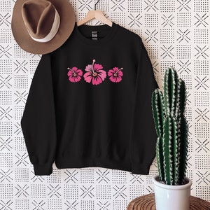 May include: Black sweatshirt with a pink hibiscus flower design. The sweatshirt is hanging on a wooden hanger against a white background with a black and white geometric pattern.
