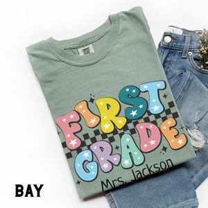 May include: Sage green t-shirt with the words "FIRST GRADE" in colorful bubble letters with star accents. The shirt includes a black and white checkered pattern and the name "Mrs. Jackson". The shirt is paired with blue denim jeans.