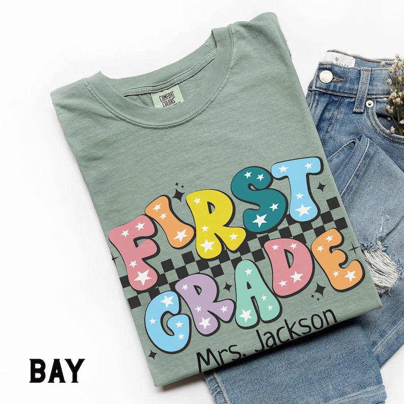 May include: Sage green t-shirt with the words "FIRST GRADE" in colorful bubble letters with star accents. The shirt includes a black and white checkered pattern and the name "Mrs. Jackson". The shirt is paired with blue denim jeans.