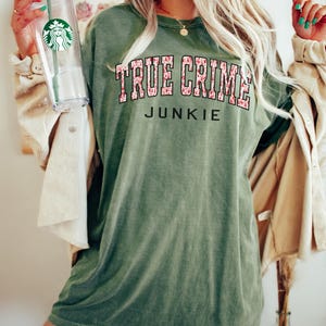 K&ouml;nnte beinhalten: Moosgr&uuml;nes T-Shirt mit dem Text "TRUE CRIME JUNKIE" in einer kr&auml;ftigen Schriftart im College-Stil. Der Text ist mit einem floralen Muster und einer schwarzen Umrandung versehen. Das Shirt hat eine lockere Passform und ist erdfarben gr&uuml;n.