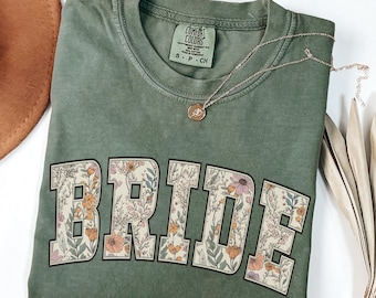 May include: A moss green t-shirt with the word "BRIDE" in large, floral-filled letters. The shirt has a crew neck and short sleeves. A gold necklace with a pendant rests on the shirt. The shirt is folded on a white surface.