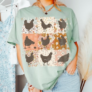 May include: A light green t-shirt with a patchwork design featuring brown silhouettes of chickens and roosters. The design includes floral patterns in shades of pink, orange, and yellow.