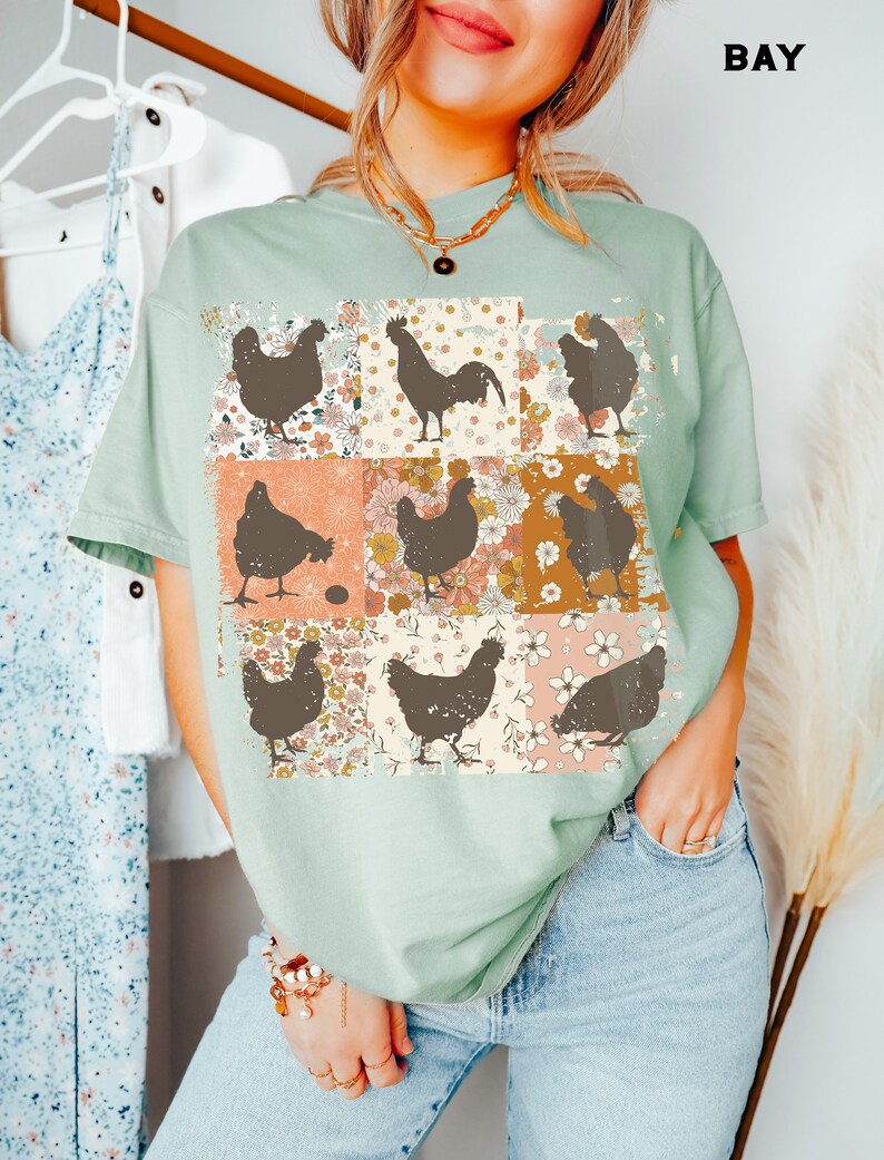 May include: A light green t-shirt with a patchwork design featuring brown silhouettes of chickens and roosters. The design includes floral patterns in shades of pink, orange, and yellow.
