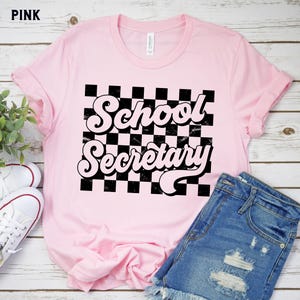 Peut inclure: T-shirt rose avec un fond &agrave; carreaux noir et blanc et le texte "School Secretary" dans une police r&eacute;tro.