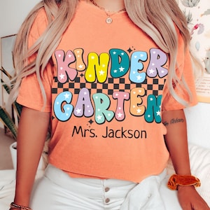 May include: Terracotta-colored t-shirt with the words "KINDER GARDEN" in a colorful, retro font. The shirt also includes the name "Mrs. Jackson" below the main text. The shirt is a short-sleeved, crew neck style.