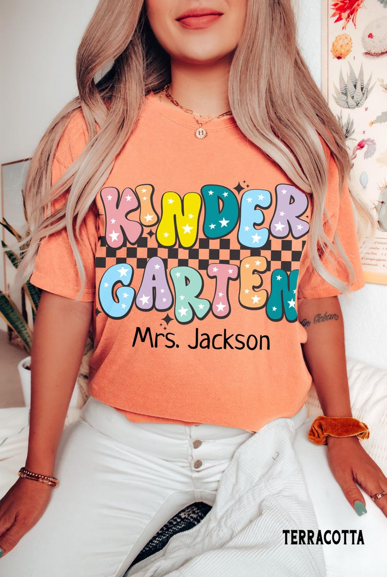 May include: Terracotta-colored t-shirt with the words "KINDER GARDEN" in a colorful, retro font. The shirt also includes the name "Mrs. Jackson" below the main text. The shirt is a short-sleeved, crew neck style.