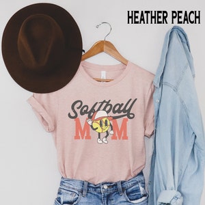 May include: Heather peach t-shirt with a softball graphic. The design features a smiling softball character wearing a cap and waving, with the words "Softball Mom" in a vintage style. A brown hat and a denim jacket are also in the image.