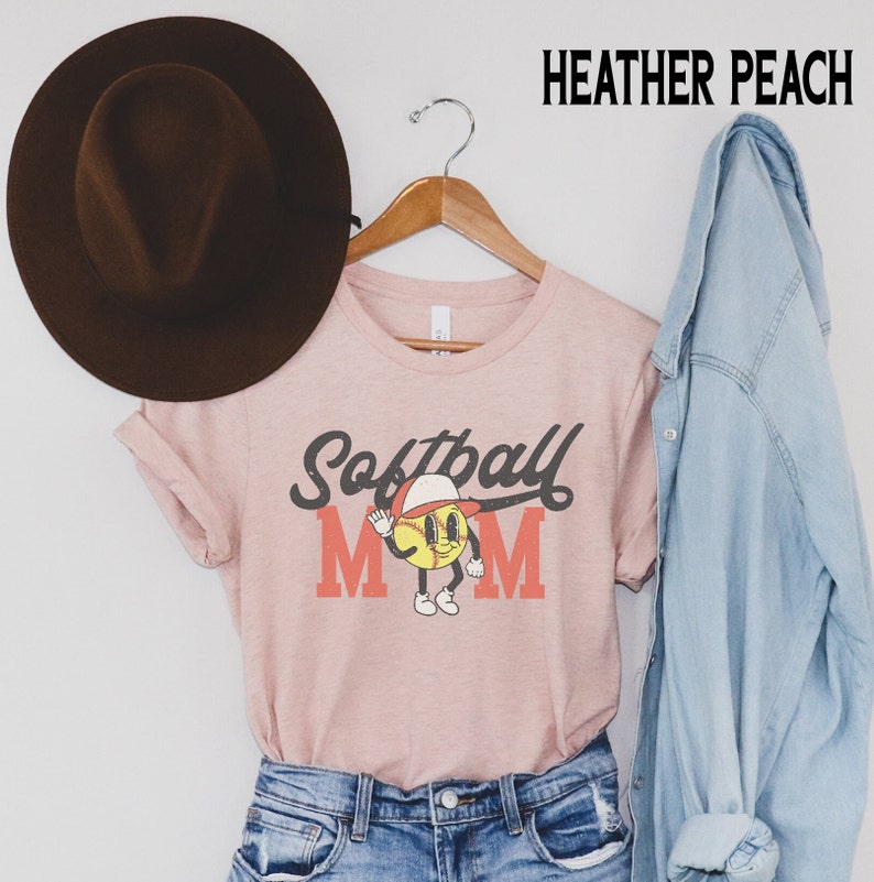 May include: Heather peach t-shirt with a softball graphic. The design features a smiling softball character wearing a cap and waving, with the words "Softball Mom" in a vintage style. A brown hat and a denim jacket are also in the image.