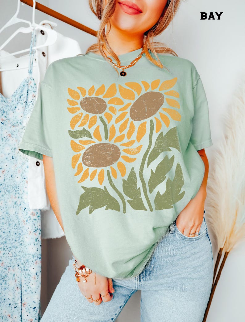 May include: A light green t-shirt with a graphic print of three stylized sunflowers in yellow, orange, and brown with green leaves. The shirt has a relaxed fit and short sleeves.
