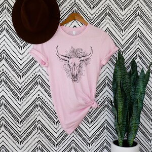 Peut inclure: Un t-shirt rose avec un motif noir et blanc de cr&acirc;ne de taureau avec des accents floraux.