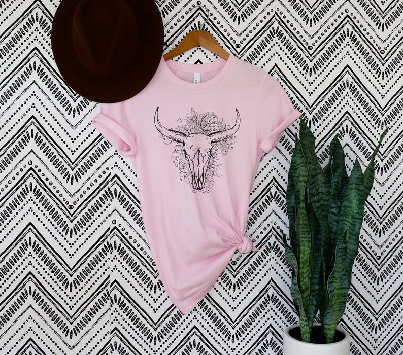 Peut inclure: Un t-shirt rose avec un motif noir et blanc de cr&acirc;ne de taureau avec des accents floraux.