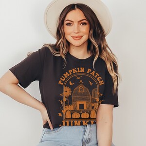 Puede incluir: Una camiseta negra con un gr&aacute;fico naranja de una granja, calabazas y el texto "Pumpkin Patch Junkie".
