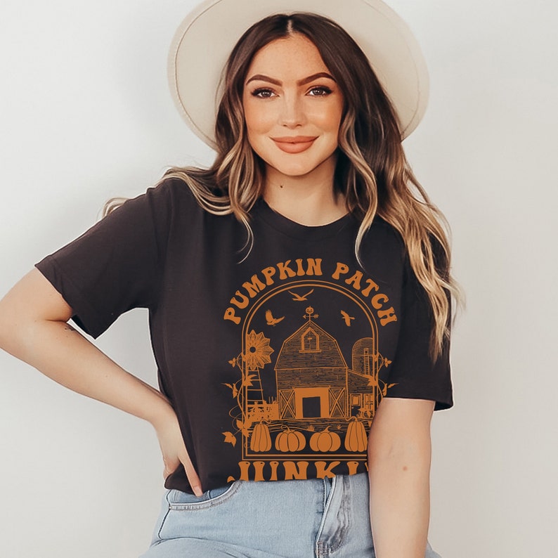 Puede incluir: Una camiseta negra con un gr&aacute;fico naranja de una granja, calabazas y el texto "Pumpkin Patch Junkie".