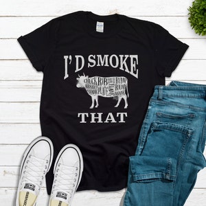 Puede incluir: Camiseta negra con un gr&aacute;fico blanco de una vaca con diferentes cortes de carne etiquetados. El texto "I'd Smoke That" est&aacute; debajo de la vaca.