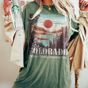 May include: Green t-shirt with a Colorado landscape graphic featuring a lake, mountains, and a red sun. The text "COLORADO Brown Family Vacation '26" is printed below the graphic. A clear Starbucks tumbler with a green straw is held in the image.