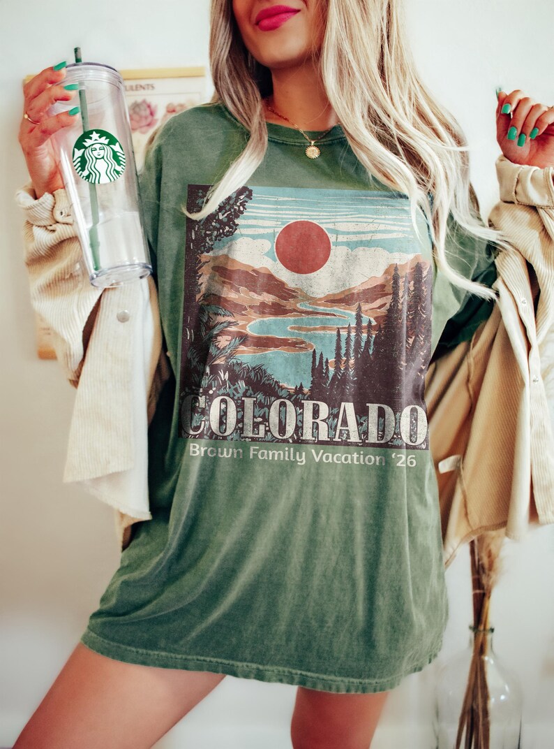 May include: Green t-shirt with a Colorado landscape graphic featuring a lake, mountains, and a red sun. The text "COLORADO Brown Family Vacation '26" is printed below the graphic. A clear Starbucks tumbler with a green straw is held in the image.