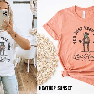 Puede incluir: Dos camisetas con una rana que lleva un sombrero de vaquero y sostiene una pistola. El texto en las camisetas dice "You just yeed your last haw". Las camisetas est&aacute;n disponibles en blanco y en color atardecer de brezo.
