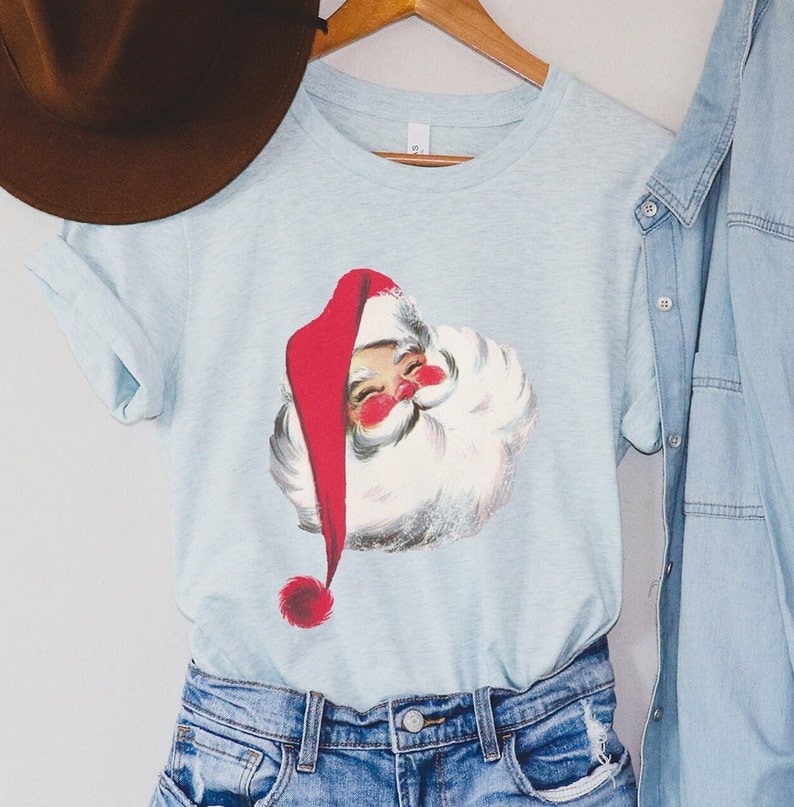 May include: A light blue t-shirt with a vintage illustration of Santa Claus's face. The illustration is in red, white, and black.