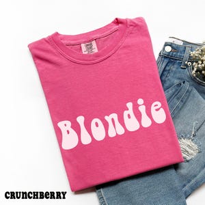 Puede incluir: Una camiseta rosa con la palabra "Blondie" en letras blancas y estilizadas. La camiseta est&aacute; doblada y colocada junto a unos vaqueros azules. La palabra "Crunchberry" est&aacute; en la parte inferior izquierda.