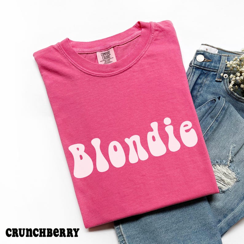 Puede incluir: Una camiseta rosa con la palabra "Blondie" en letras blancas y estilizadas. La camiseta est&aacute; doblada y colocada junto a unos vaqueros azules. La palabra "Crunchberry" est&aacute; en la parte inferior izquierda.