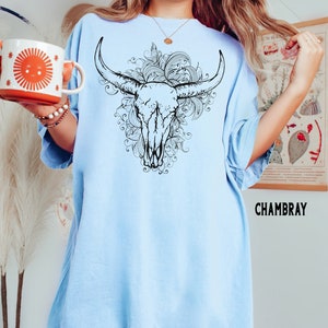 K&ouml;nnte beinhalten: Ein hellblaues Oversize-T-Shirt mit einer schwarzen und wei&szlig;en Grafik eines Bullensch&auml;dels mit floralen Details. Der Text "CHAMBRAY" ist in der rechten unteren Ecke des Shirts gedruckt.