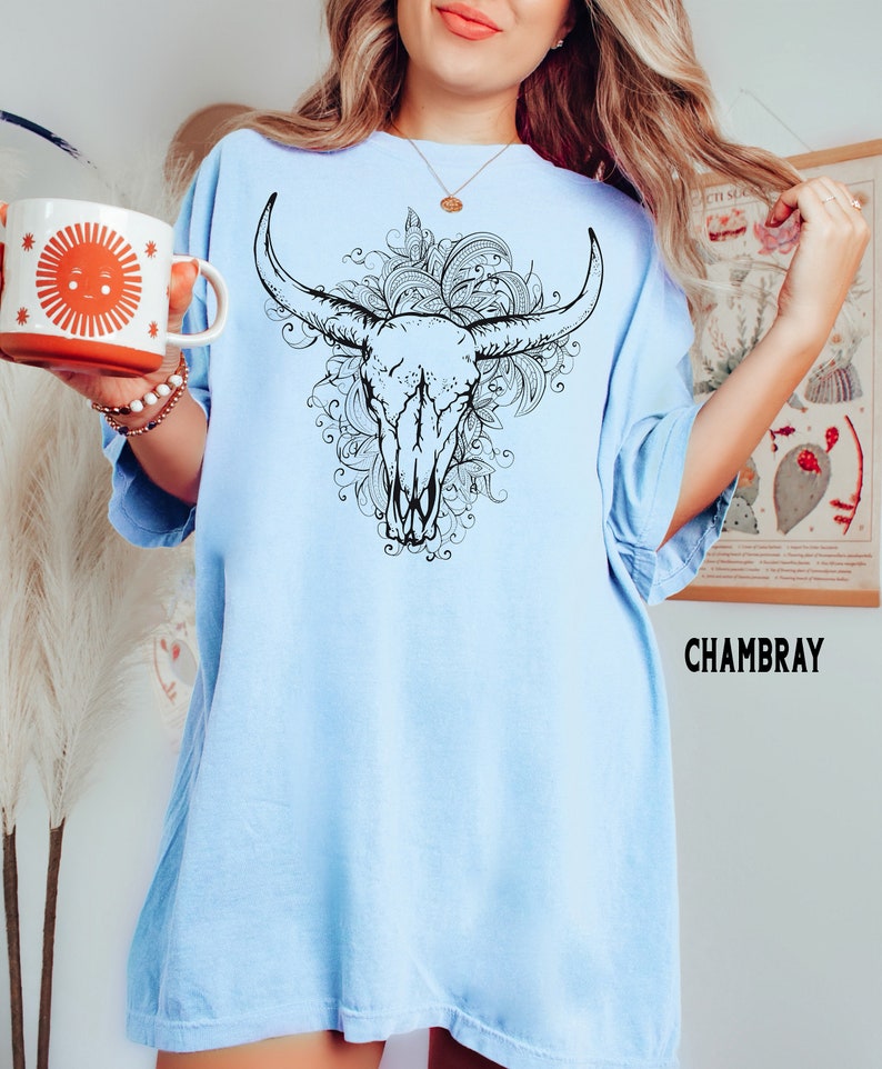 K&ouml;nnte beinhalten: Ein hellblaues Oversize-T-Shirt mit einer schwarzen und wei&szlig;en Grafik eines Bullensch&auml;dels mit floralen Details. Der Text "CHAMBRAY" ist in der rechten unteren Ecke des Shirts gedruckt.