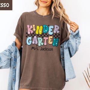 May include: Espresso-colored t-shirt with the word "KINDERGARTEN" in colorful bubble letters, a checkered pattern, and the name "Mrs. Jackson" below. The shirt is worn with a denim jacket.