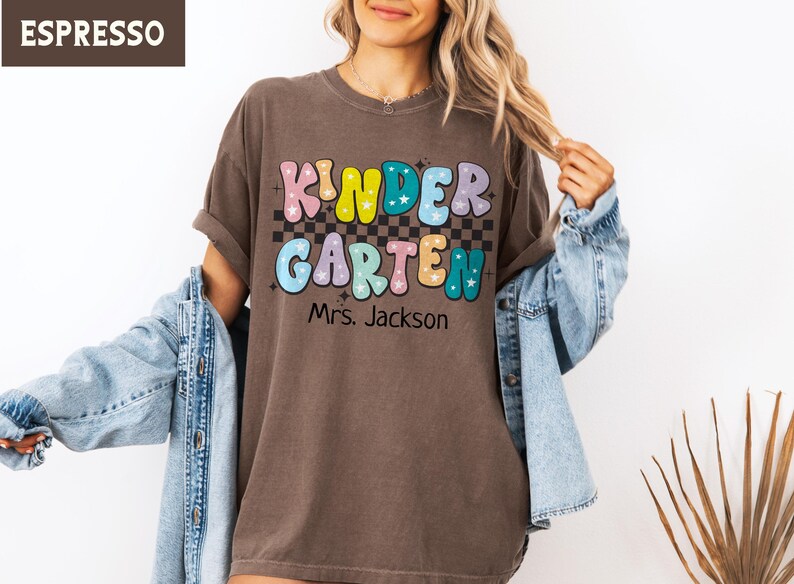 May include: Espresso-colored t-shirt with the word "KINDERGARTEN" in colorful bubble letters, a checkered pattern, and the name "Mrs. Jackson" below. The shirt is worn with a denim jacket.