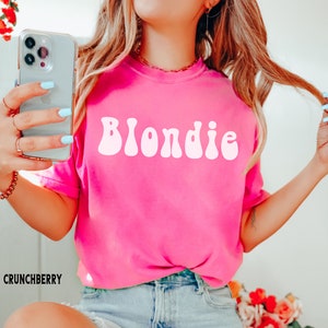 以下が含まれることがあります： ピンクのTシャツに、レトロなスタイルの白い文字で「Blondie」と書かれています。このTシャツは、長いブロンドの髪をした人が着ています。