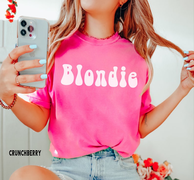 以下が含まれることがあります： ピンクのTシャツに、レトロなスタイルの白い文字で「Blondie」と書かれています。このTシャツは、長いブロンドの髪をした人が着ています。