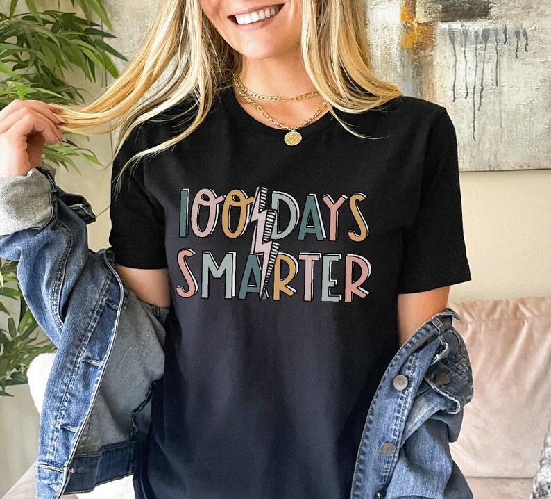 Pode incluir: Camiseta preta com o texto "100 Days Smarter" em letras coloridas com um gr&aacute;fico de raio.