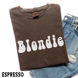 Puede incluir: Camiseta color espresso con la palabra "Blondie" en letras blancas de estilo retro. La camiseta est&aacute; doblada y colocada junto a un par de vaqueros de mezclilla azules. La camiseta tiene cuello redondo y una etiqueta en el cuello.