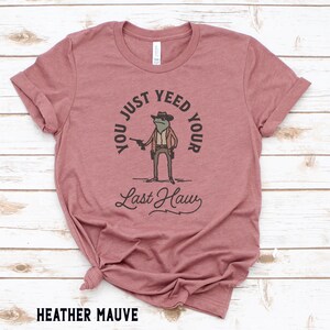 Puede incluir: Camiseta de color malva jaspeado con un dibujo de una rana de dibujos animados que lleva un sombrero de vaquero y sostiene una pistola. El texto de la camiseta dice "You just yeed your last haw".