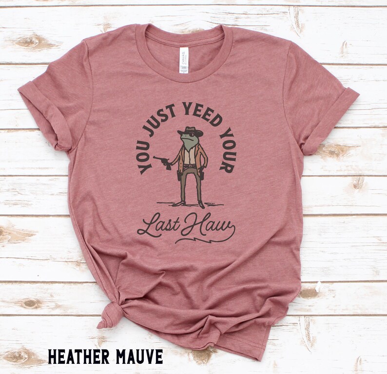 Puede incluir: Camiseta de color malva jaspeado con un dibujo de una rana de dibujos animados que lleva un sombrero de vaquero y sostiene una pistola. El texto de la camiseta dice "You just yeed your last haw".