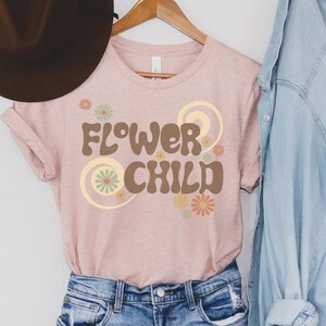 Puede incluir: Una camiseta rosa claro con la frase "FLOWER CHILD" en marr&oacute;n, con letras de estilo retro. El dise&ntilde;o incluye detalles florales en tonos naranja, verde y amarillo. La camiseta est&aacute; colgada en una percha de madera.