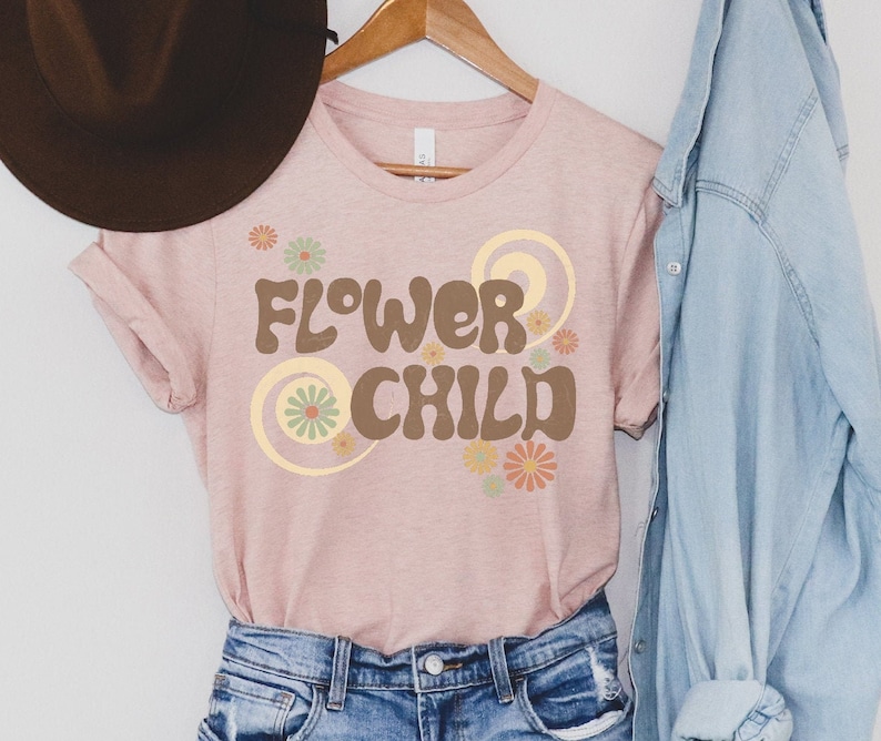 Puede incluir: Una camiseta rosa claro con la frase "FLOWER CHILD" en marr&oacute;n, con letras de estilo retro. El dise&ntilde;o incluye detalles florales en tonos naranja, verde y amarillo. La camiseta est&aacute; colgada en una percha de madera.