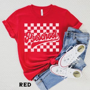 Pu&ograve; includere: T-shirt rosso con la scritta "Preschool" in un font retr&ograve; su un motivo a scacchi bianchi e rossi. La maglietta &egrave; abbinata a jeans blu e scarpe da ginnastica bianche.