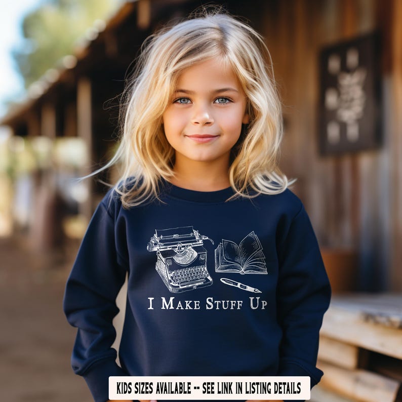 Puede incluir: Un ni&ntilde;o lleva una sudadera azul marino con un gr&aacute;fico blanco de una m&aacute;quina de escribir, un libro abierto y un bol&iacute;grafo. El texto "I MAKE STUFF UP" est&aacute; impreso debajo. La sudadera forma parte de una colecci&oacute;n infantil.