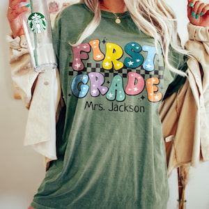 May include: Olive green t-shirt with the words "FIRST GRADE Mrs. Jackson" in colorful, retro-style lettering. The shirt has a checkerboard pattern behind the text and star accents. A clear Starbucks tumbler is held in the left hand.