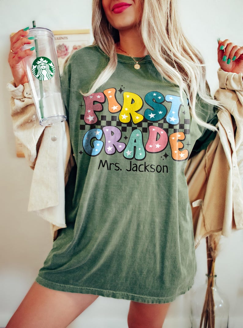 May include: Olive green t-shirt with the words "FIRST GRADE Mrs. Jackson" in colorful, retro-style lettering. The shirt has a checkerboard pattern behind the text and star accents. A clear Starbucks tumbler is held in the left hand.