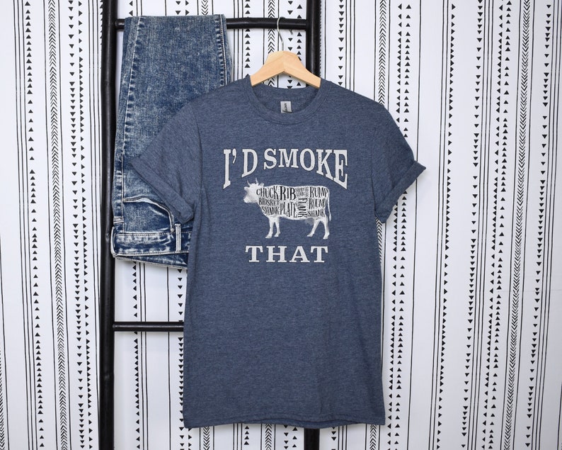 Puede incluir: Una camiseta azul oscuro con un gr&aacute;fico blanco de una vaca con diferentes cortes de carne etiquetados. El texto "I'd Smoke That" est&aacute; impreso debajo de la vaca.