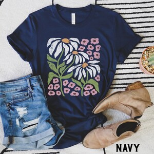 K&ouml;nnte beinhalten: Ein marineblaues T-Shirt mit einem wei&szlig;en G&auml;nsebl&uuml;mchen-Blumendruck. Der Druck zeigt zwei gro&szlig;e G&auml;nsebl&uuml;mchen, die von kleineren rosa Bl&uuml;ten umgeben sind. Das Shirt hat einen Rundhalsausschnitt und kurze &Auml;rmel. Das Wort "NAVY" ist in Wei&szlig; am unteren Rand des Bildes aufgedruckt.