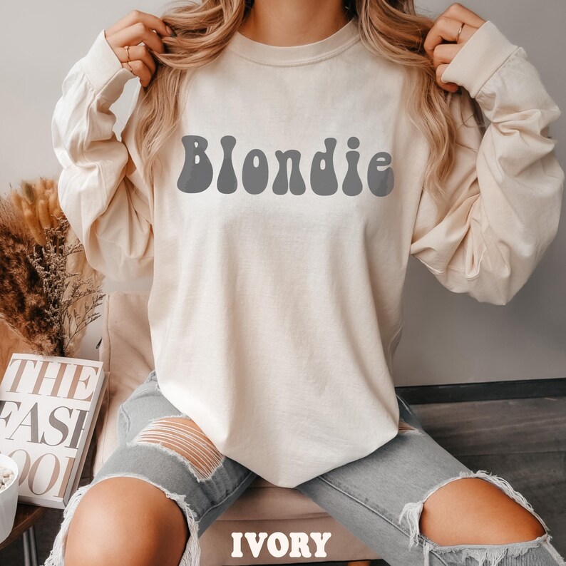 Puede incluir: Camiseta de manga larga color marfil con la palabra "Blondie" en gris, con letras de estilo retro. La camiseta es de color neutro y tiene un corte holgado. La palabra "IVORY" est&aacute; impresa en la parte inferior de la imagen.