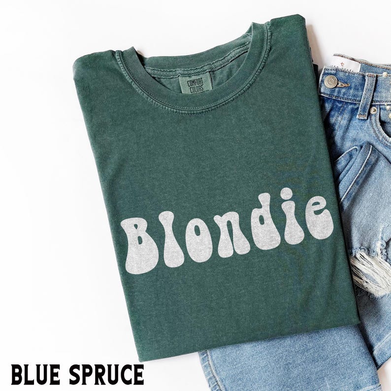Puede incluir: Una camiseta azul abeto con la palabra "Blondie" en blanco, con letras de estilo retro. La camiseta est&aacute; doblada y colocada junto a unos pantalones cortos vaqueros. La etiqueta dice "Comfort Colors".