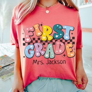 May include: A coral-colored t-shirt with the words "FIRST GRADE" in colorful, retro bubble letters. The text is over a black and white checkered pattern. Below the text is "Mrs. Jackson". The word "WATERMELON" is at the bottom.