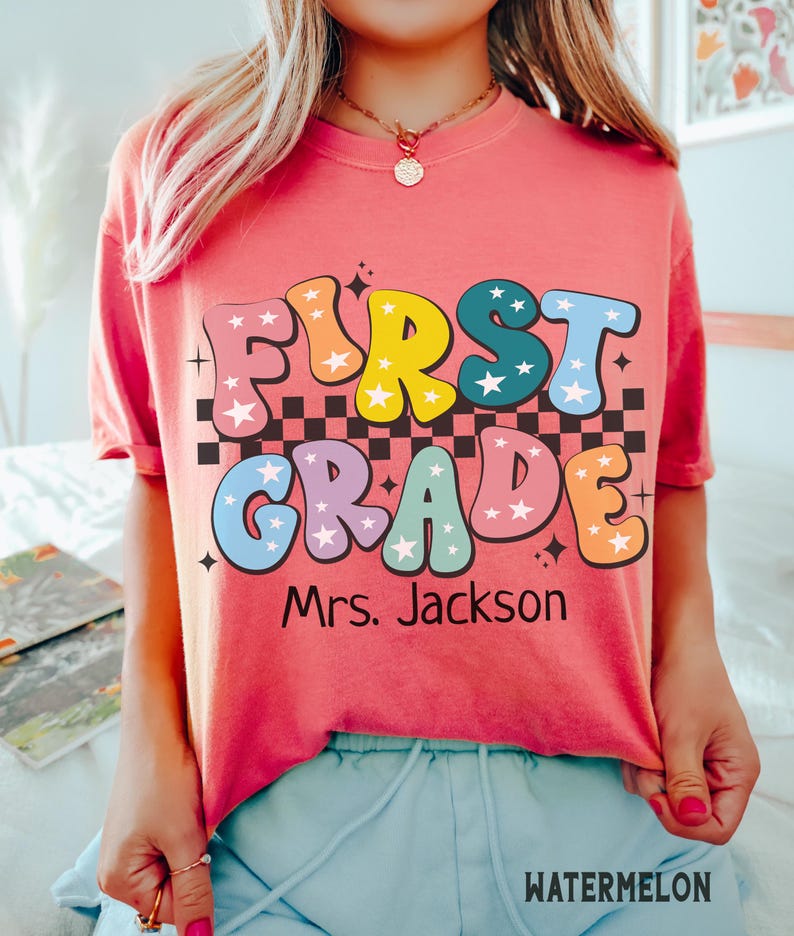 May include: A coral-colored t-shirt with the words "FIRST GRADE" in colorful, retro bubble letters. The text is over a black and white checkered pattern. Below the text is "Mrs. Jackson". The word "WATERMELON" is at the bottom.