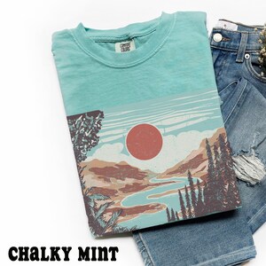 May include: A Chalky Mint colored t-shirt with a landscape graphic featuring a red sun, river, and trees. The shirt is folded on top of a pair of blue denim shorts.