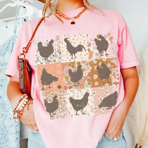 May include: Pink t-shirt with a patchwork design featuring brown silhouettes of chickens and a rooster. The patchwork design includes floral patterns in shades of pink, orange, and yellow.